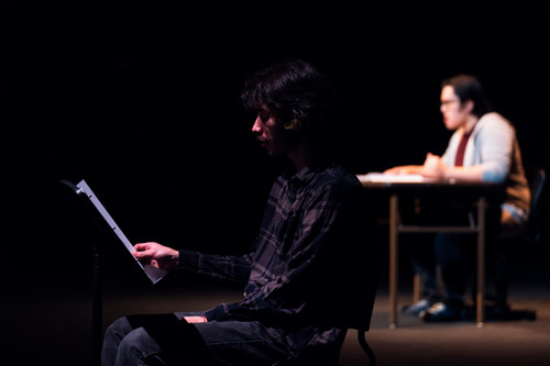 A man reads from a script on a music stand with another actor in the distanceChase Keelin reads Stage Directions, Bennett Buchholz as Silas