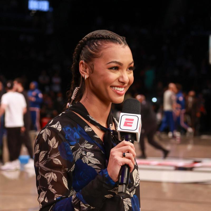 Malika Andrews, a black woman smiling on a NBA basketball court with a microphone in her hand