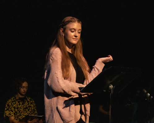 A blond girl in a pink sweater reads from a music stand